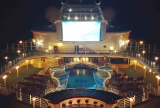 A luxury cruise ship deck featuring a pool surrounded by lounge chairs and warm ambient lighting. A large screen overlooks the area, showing an image against a dark night sky.