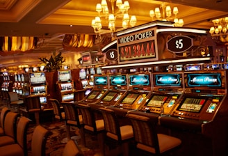 A casino floor featuring rows of brightly lit video poker machines. The area is illuminated by elegant chandeliers, with a display board showing a large sum of money, suggesting a possible jackpot. The rich decor includes luxurious carpets and comfortable seating, adding to the opulent atmosphere.