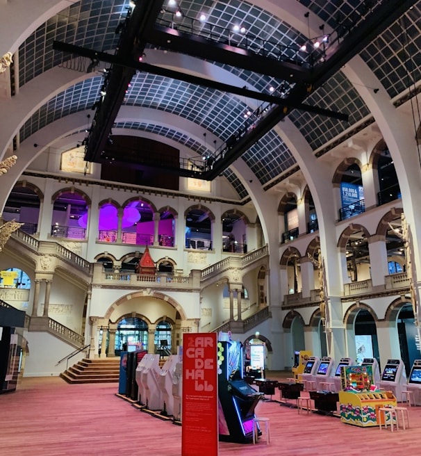 The interior of a large, ornately designed building with arched ceilings and intricate architectural details. The space features gaming machines lined up along the floor, under a high ceiling with a grid pattern. Lighting in purple and white tones enhances the ambiance.