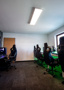 A well-lit gaming room features multiple gaming stations with high-backed chairs and monitors. The room has a minimalist setup with green and black color tones. Large posters adorn the walls, and the room appears clean and organized.