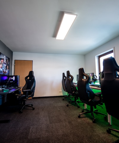 A well-lit gaming room features multiple gaming stations with high-backed chairs and monitors. The room has a minimalist setup with green and black color tones. Large posters adorn the walls, and the room appears clean and organized.