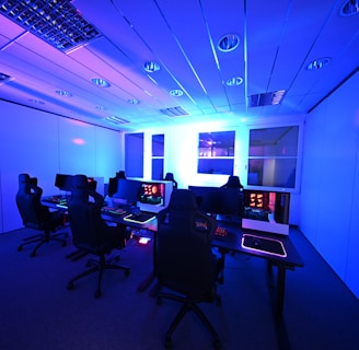 A modern gaming setup with multiple high-backed gaming chairs and monitors arranged in a room with a cool blue lighting. The computers have illuminated keyboards and glowing screens, creating a high-tech ambiance.