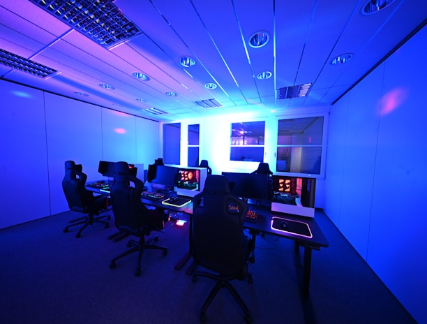 A modern gaming setup with multiple high-backed gaming chairs and monitors arranged in a room with a cool blue lighting. The computers have illuminated keyboards and glowing screens, creating a high-tech ambiance.