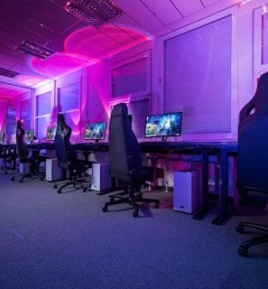 A row of gaming computers with large monitors and racing-style chairs is set up along the interior of a modern room. The space is illuminated with vibrant purple and blue LED lighting, creating a futuristic atmosphere. The room is organized and clean, suggesting a professional gaming or esports environment.