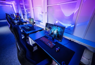A row of modern gaming setups is arranged neatly along a window-lined wall. Each station includes a high-resolution monitor displaying vivid graphics, a mechanical keyboard with colored lighting, a gaming mouse, and an ergonomic chair. The room is illuminated with ambient blue and purple lights, creating a sleek and futuristic atmosphere.