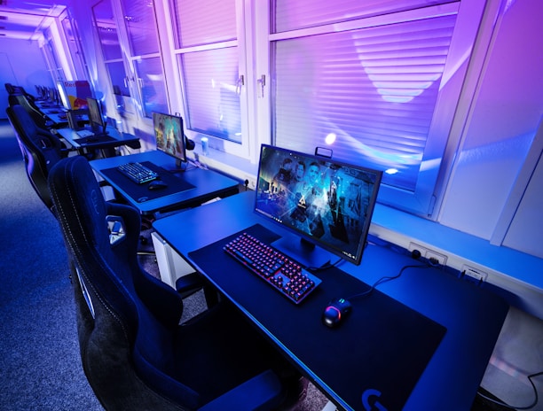 A row of modern gaming setups is arranged neatly along a window-lined wall. Each station includes a high-resolution monitor displaying vivid graphics, a mechanical keyboard with colored lighting, a gaming mouse, and an ergonomic chair. The room is illuminated with ambient blue and purple lights, creating a sleek and futuristic atmosphere.