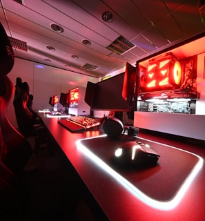 A modern gaming setup featuring several high-end computers with illuminated red interior lighting. The room is equipped with gaming chairs exhibiting a sleek black and red design. Multiple monitors are arranged on a long desk, and peripheral devices like keyboards and mice are uniformly placed, reflecting an ambiance ideal for gaming or competitive play.