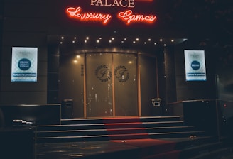 A neon-lit casino entrance with a red carpet leading up to large double doors. The sign reads 'Fortuna Palace Luxury Games' in bright red and white neon lights. The dimly lit atmosphere suggests a night-time setting. There are two digital displays on either side of the entrance showing promotional content.