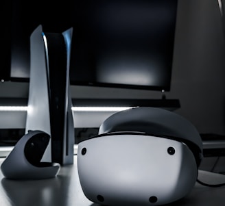 A sleek virtual reality headset sits on a reflective surface beside a gaming console and its controller. The background features a large dark monitor screen, creating a modern and futuristic setting. The lighting is subdued, highlighting the glossy surfaces of the electronic devices.