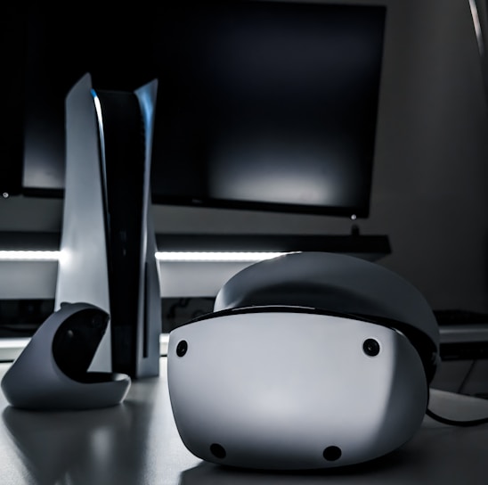 A sleek virtual reality headset sits on a reflective surface beside a gaming console and its controller. The background features a large dark monitor screen, creating a modern and futuristic setting. The lighting is subdued, highlighting the glossy surfaces of the electronic devices.