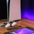 A sleek, modern setup featuring a gaming console with two controllers on a wooden surface. Nearby are a smartphone, smartwatch, tablet with a vivid screen, wireless earbuds, and a stylus. The background is illuminated with a soft purple glow.
