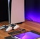 A sleek, modern setup featuring a gaming console with two controllers on a wooden surface. Nearby are a smartphone, smartwatch, tablet with a vivid screen, wireless earbuds, and a stylus. The background is illuminated with a soft purple glow.